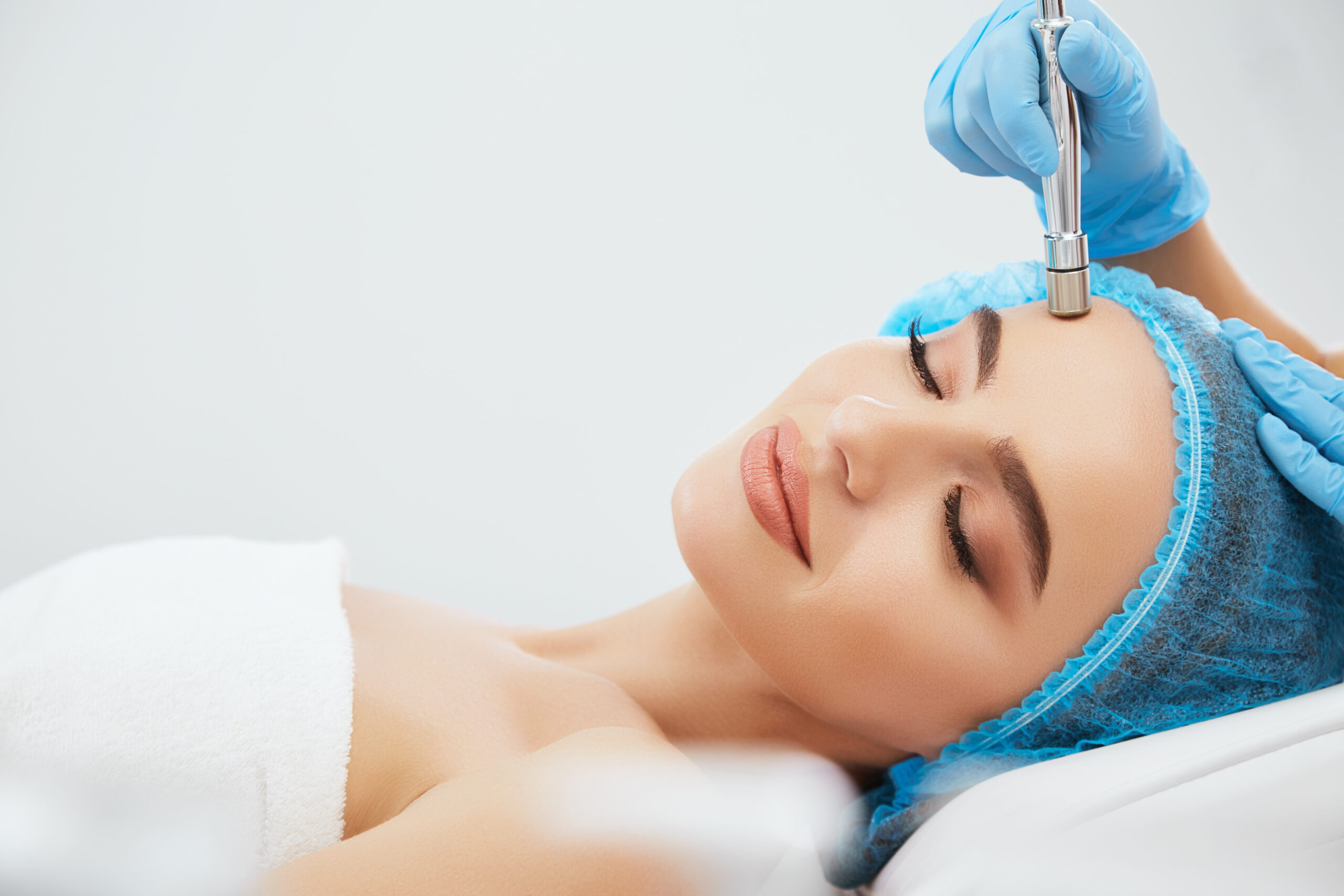 closeup smiling woman blue cap lying couch cosmetological clinic with closed eyes doctor s hands blue gloves doing procedure microdermabrasia her face scaled